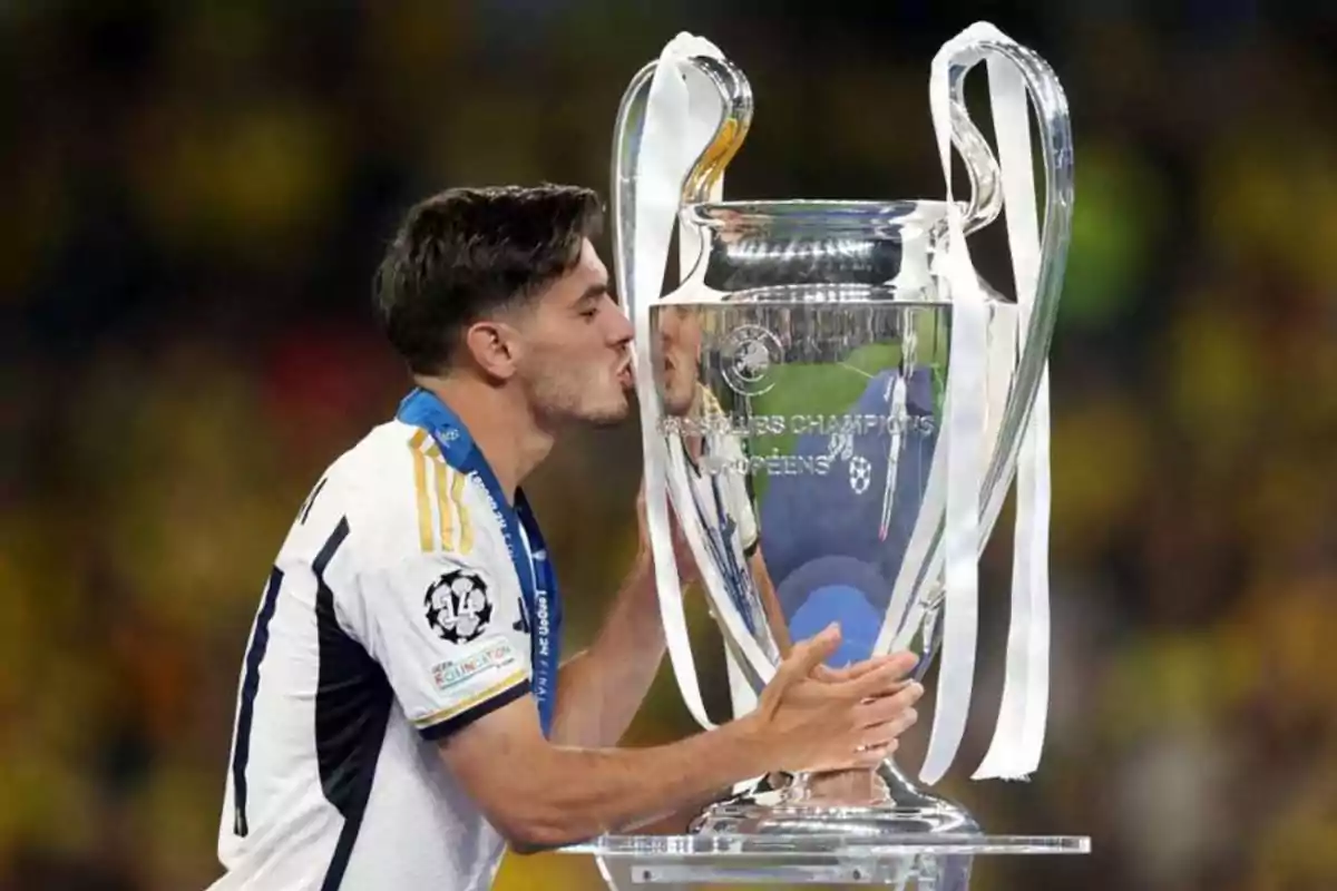 Un jugador de fútbol besa un trofeo grande y plateado mientras lleva una medalla alrededor del cuello.