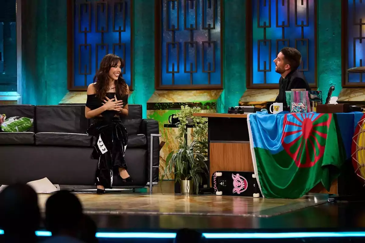 Una mujer sonriente está sentada en un sofá negro en un set de televisión, mientras un hombre la entrevista desde un escritorio decorado con una bandera colorida y varios objetos.