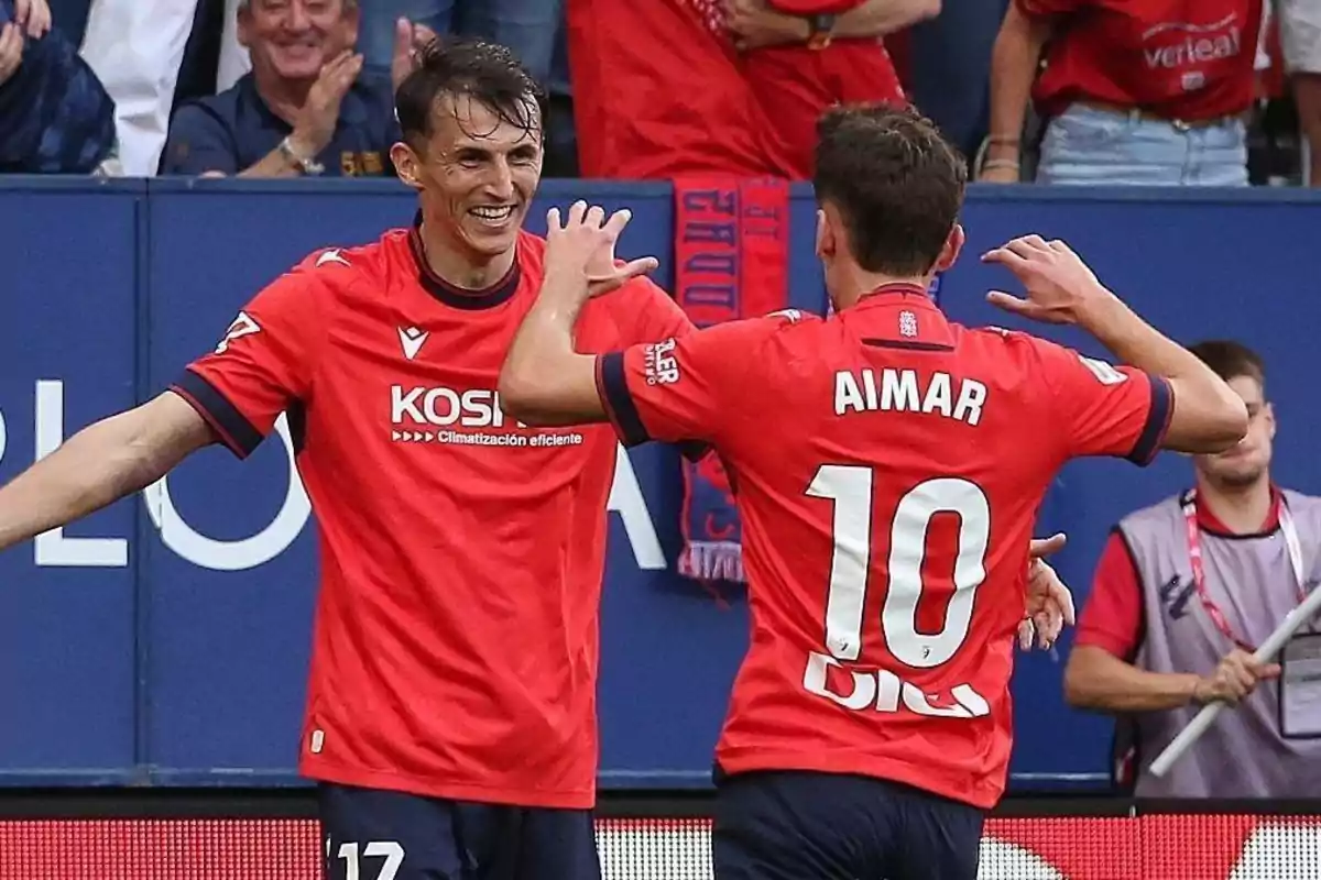 Dos futbolistas con uniforme rojo celebran un gol en un estadio mientras el público aplaude en el fondo Dos futbolistas con uniforme rojo celebran un gol en un estadio mientras el público aplaude en el fondo
