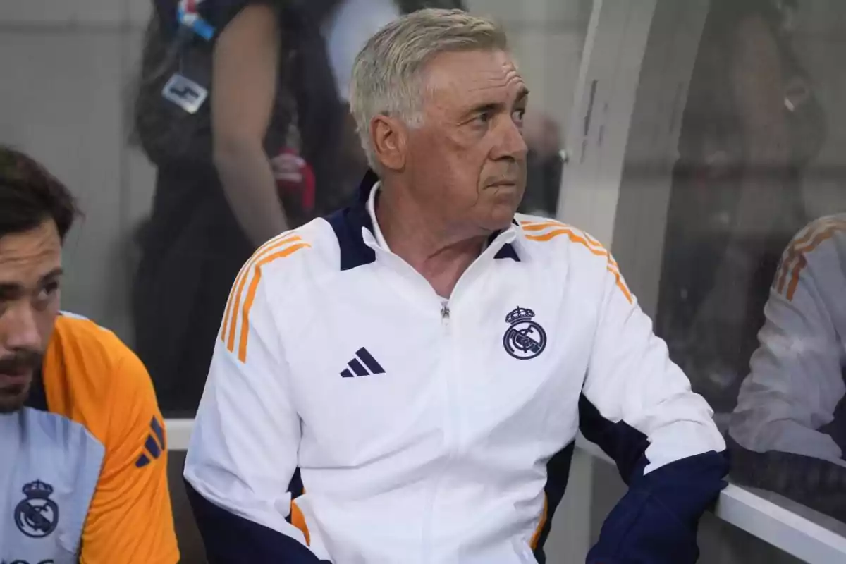 Un hombre con una chaqueta blanca y detalles en azul y naranja, con el logo del Real Madrid, sentado en un banquillo.