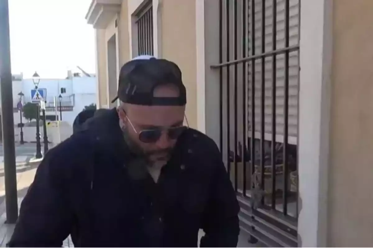 Un hombre con gorra y gafas de sol camina por la calle junto a un edificio con rejas en las ventanas. Un hombre con gorra y gafas de sol camina por la calle junto a un edificio con rejas en las ventanas.