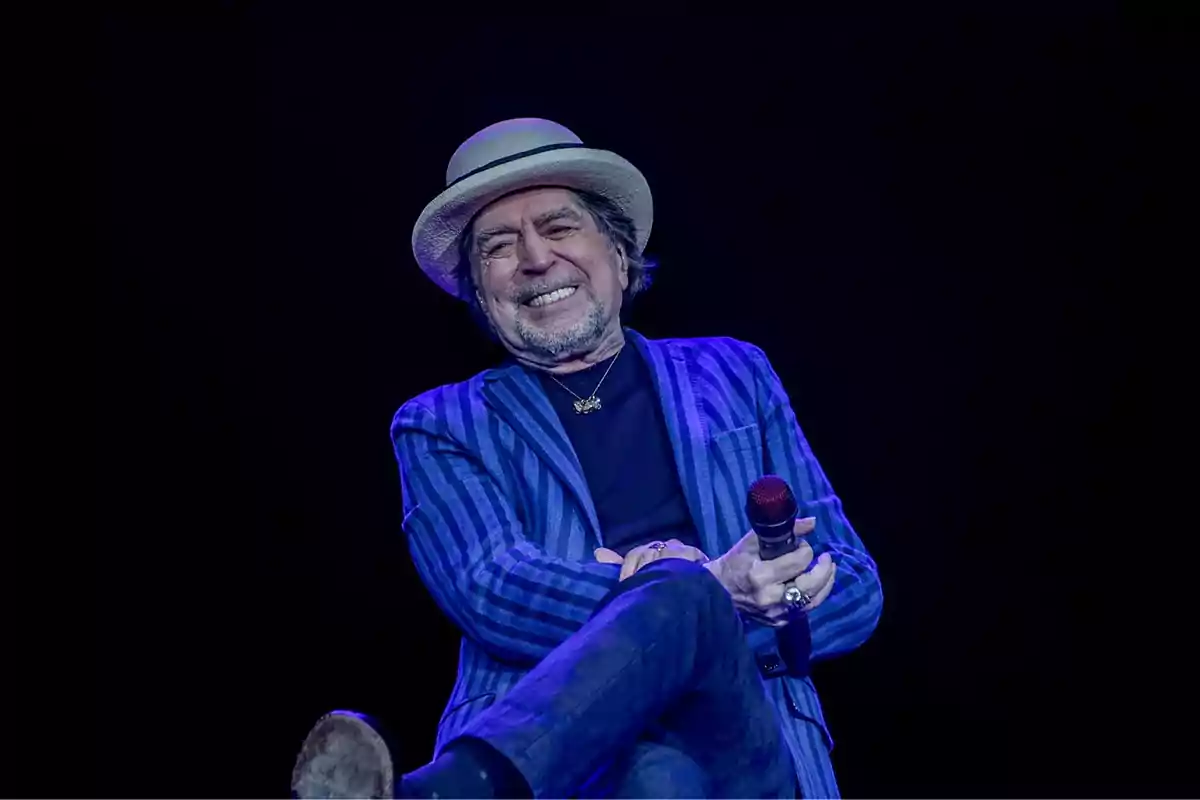 Hombre mayor con sombrero y chaqueta a rayas sonriendo mientras sostiene un micrófono en un escenario oscuro. Hombre mayor con sombrero y chaqueta a rayas sonriendo mientras sostiene un micrófono en un escenario oscuro.