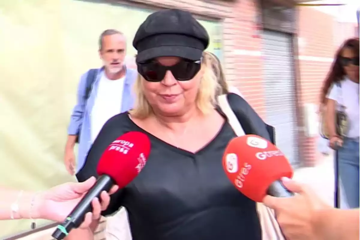 Una mujer con gafas de sol y gorra negra es entrevistada por varios medios de comunicación que sostienen micrófonos rojos. Una mujer con gafas de sol y gorra negra es entrevistada por varios medios de comunicación que sostienen micrófonos rojos.