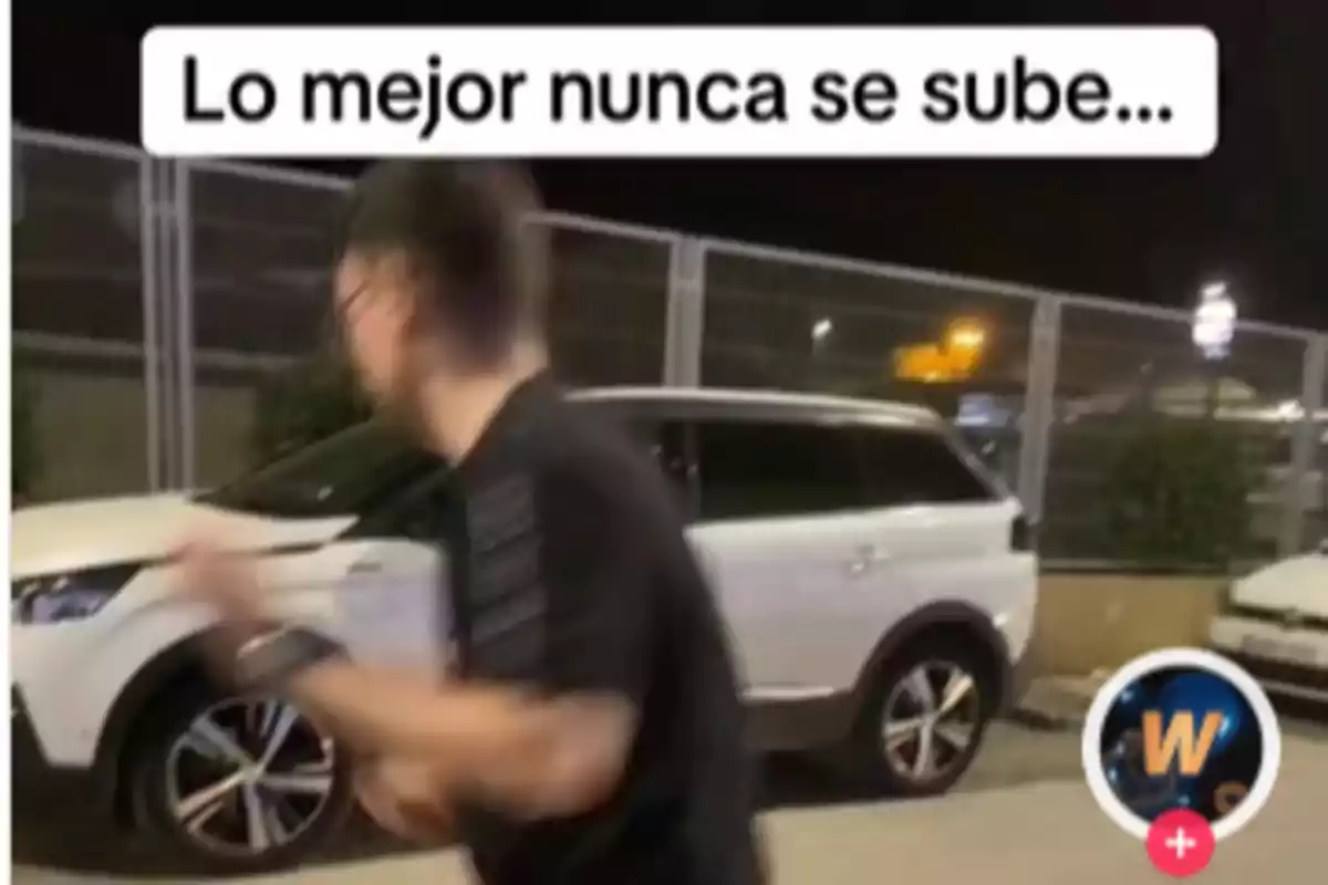 Un hombre desenfocado camina rápidamente frente a un coche blanco estacionado en un área cercada por la noche, con un texto en la parte superior que dice "Lo mejor nunca se sube".