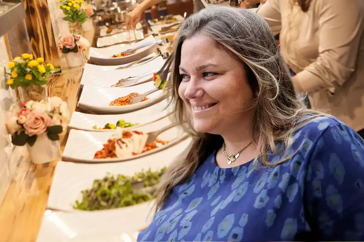 Una mujer sonriente con una blusa azul está frente a una mesa de buffet con platos de comida y decoraciones florales.