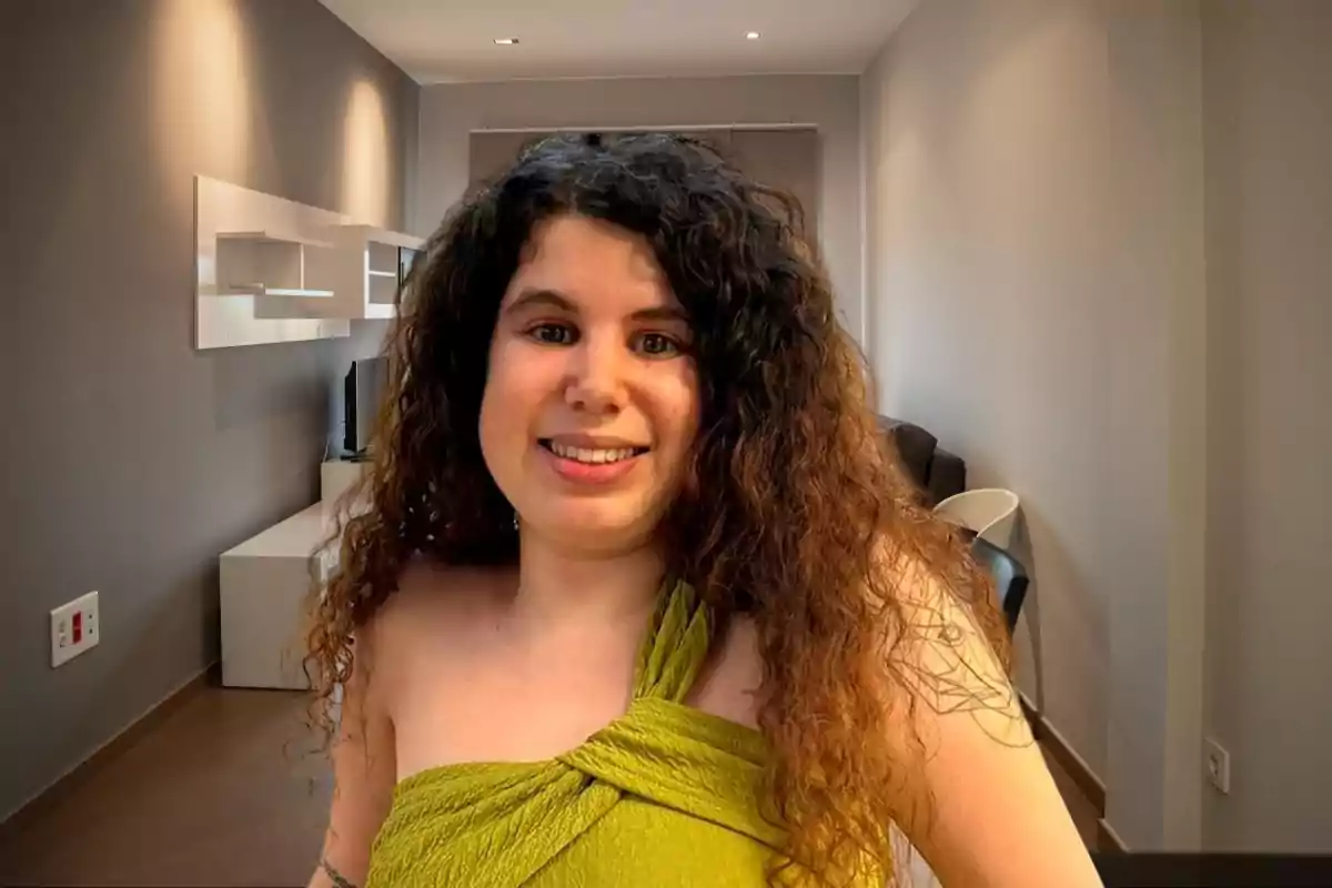 Una mujer con cabello rizado y largo, vestida con una blusa verde, sonríe mientras está en una habitación moderna con paredes grises y muebles minimalistas.