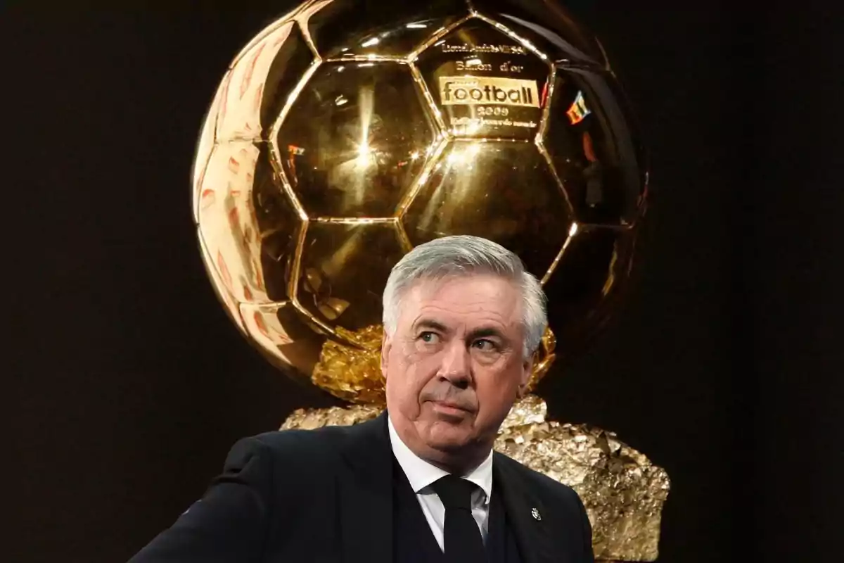 Un hombre de cabello canoso y traje oscuro está frente a un trofeo dorado con forma de balón de fútbol.