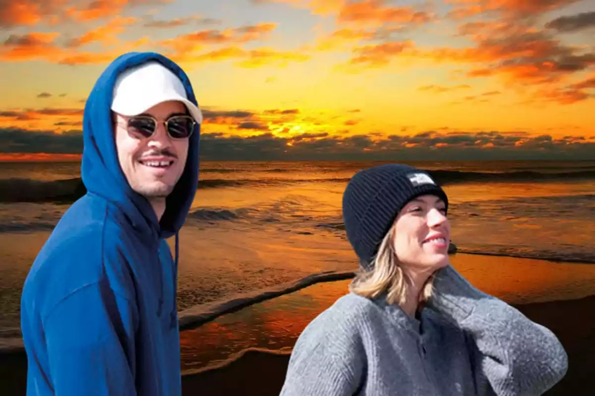 Dos personas sonrientes, una con sudadera azul y gorra blanca y otra con suéter gris y gorro negro, posan frente a un atardecer en la playa con el cielo lleno de nubes anaranjadas y el mar en calma.