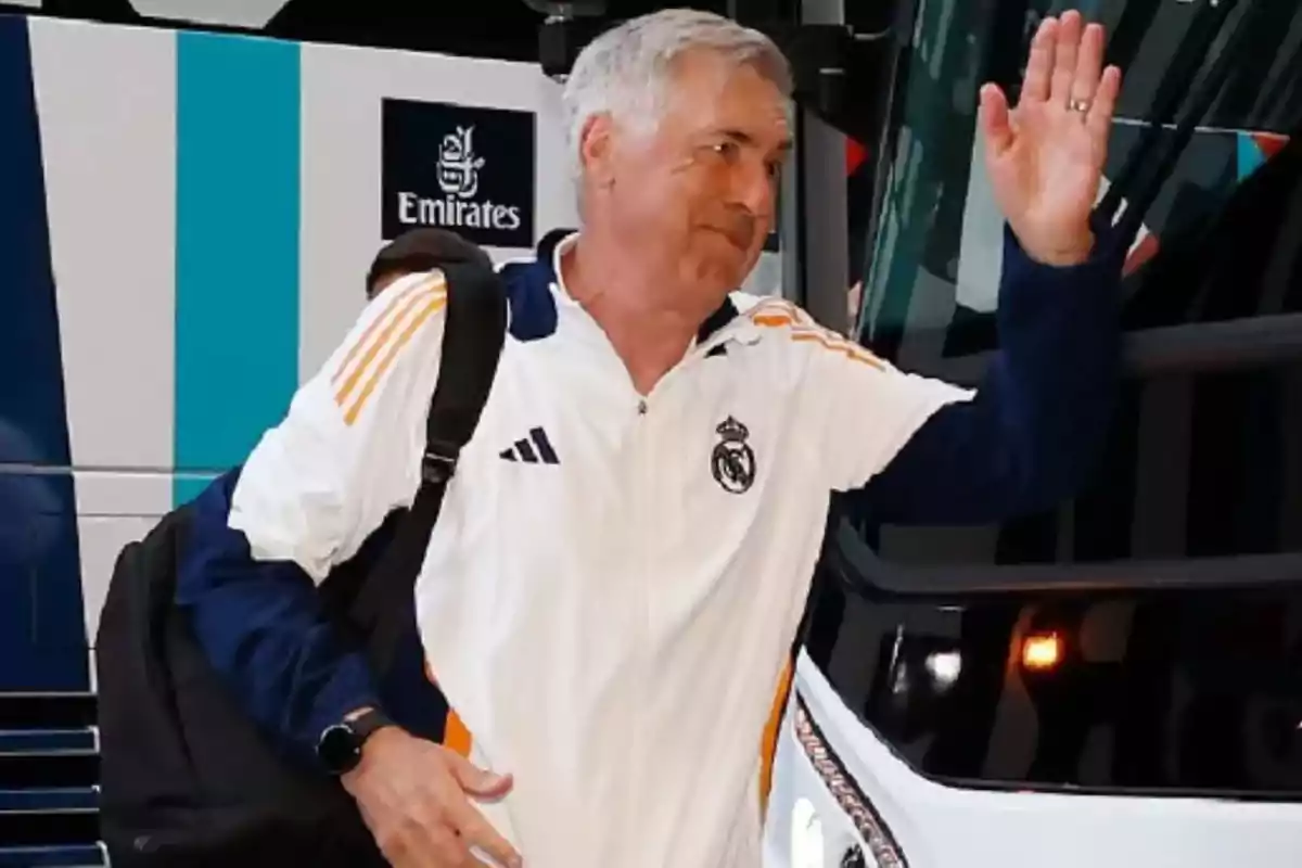 Un hombre con una chaqueta deportiva blanca y azul con el logo del Real Madrid saluda mientras se encuentra frente a un autobús.