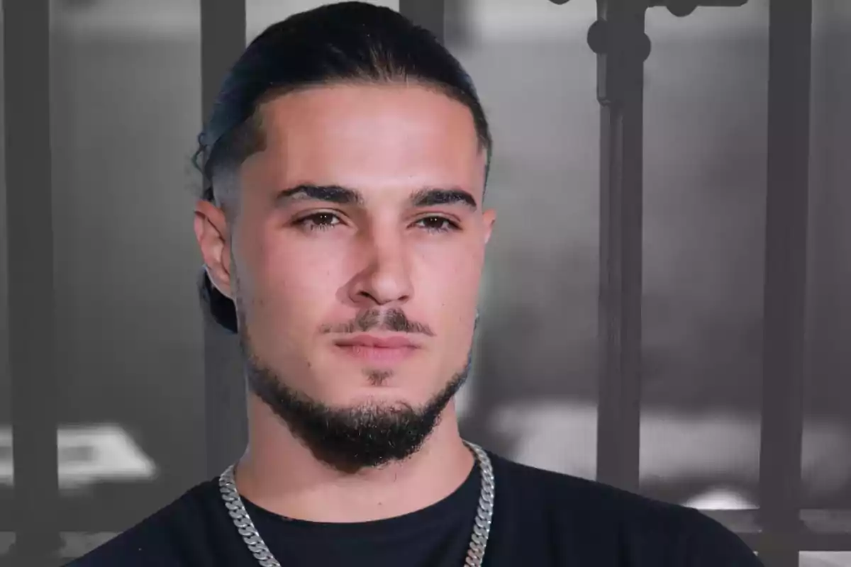 Un hombre con barba y cabello recogido mira hacia adelante con una expresión seria, lleva una cadena plateada y está frente a un fondo desenfocado.