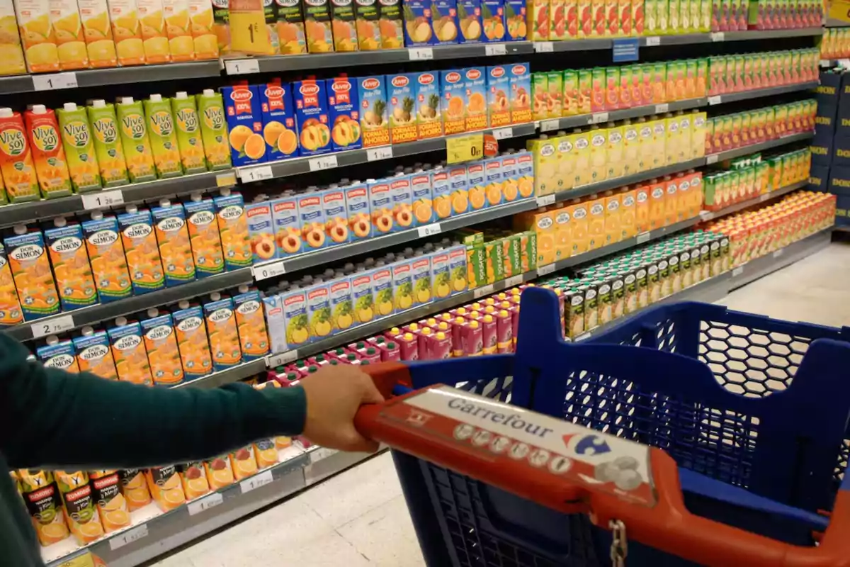 Una persona empuja un carrito de compras en un supermercado, frente a una estantería llena de diferentes marcas y tipos de jugos envasados. Una persona empuja un carrito de compras en un supermercado, frente a una estantería llena de diferentes marcas y tipos de jugos envasados.
