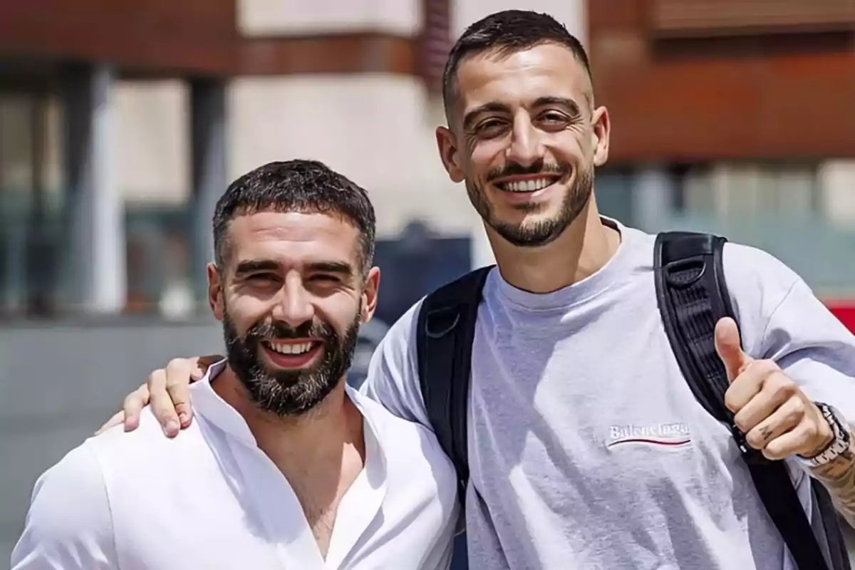 Dos hombres sonrientes posando juntos al aire libre, uno de ellos con una camiseta gris y mochila, y el otro con una camisa blanca.