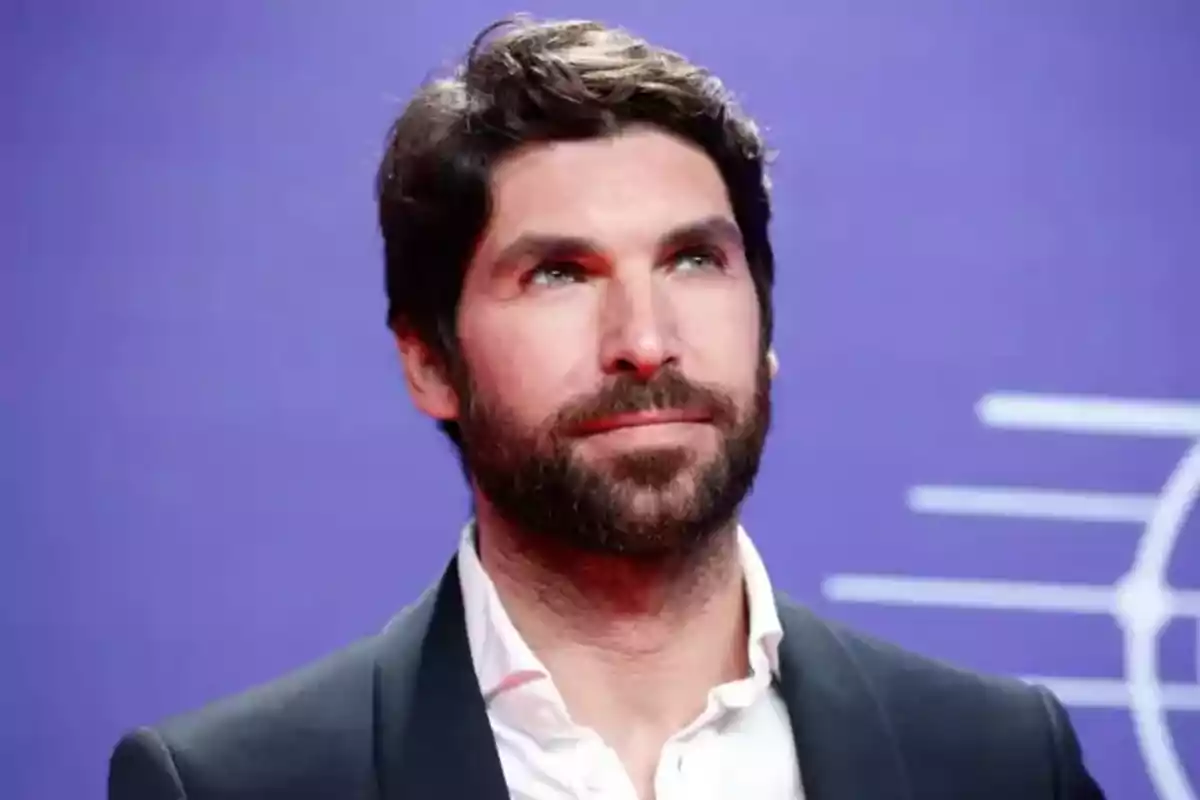 Un hombre con barba y cabello oscuro lleva una camisa blanca y un saco oscuro, posando frente a un fondo morado. Un hombre con barba y cabello oscuro lleva una camisa blanca y un saco oscuro, posando frente a un fondo morado.