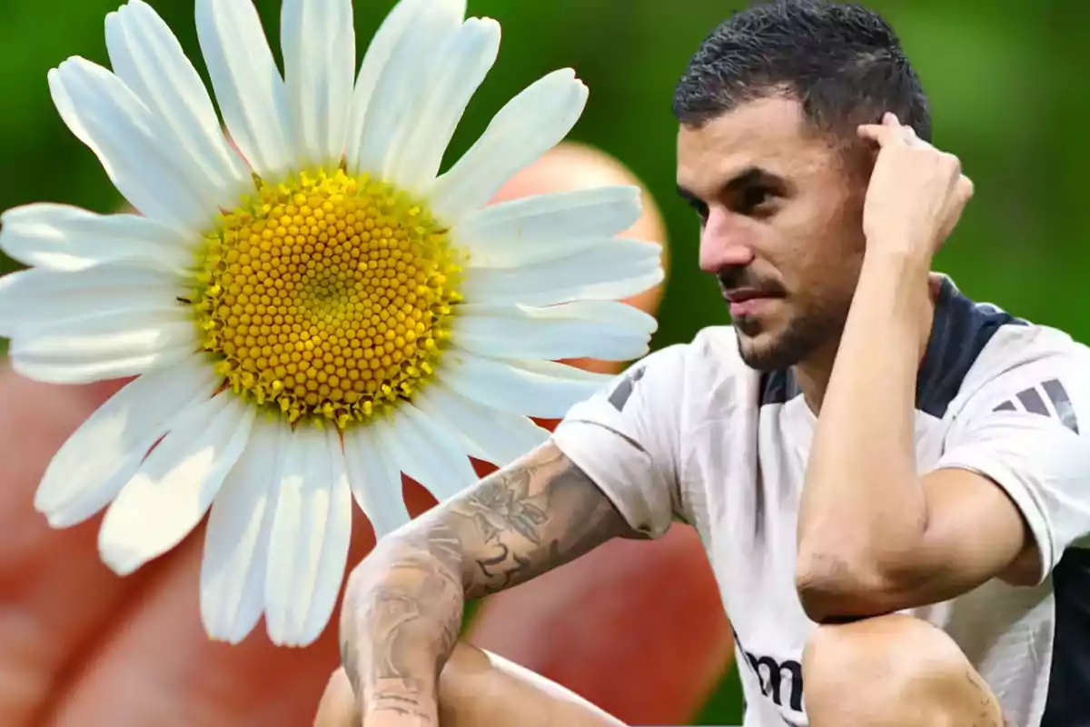 Un hombre con tatuajes en el brazo y una camiseta blanca está sentado y pensativo, con una flor blanca y amarilla en primer plano.