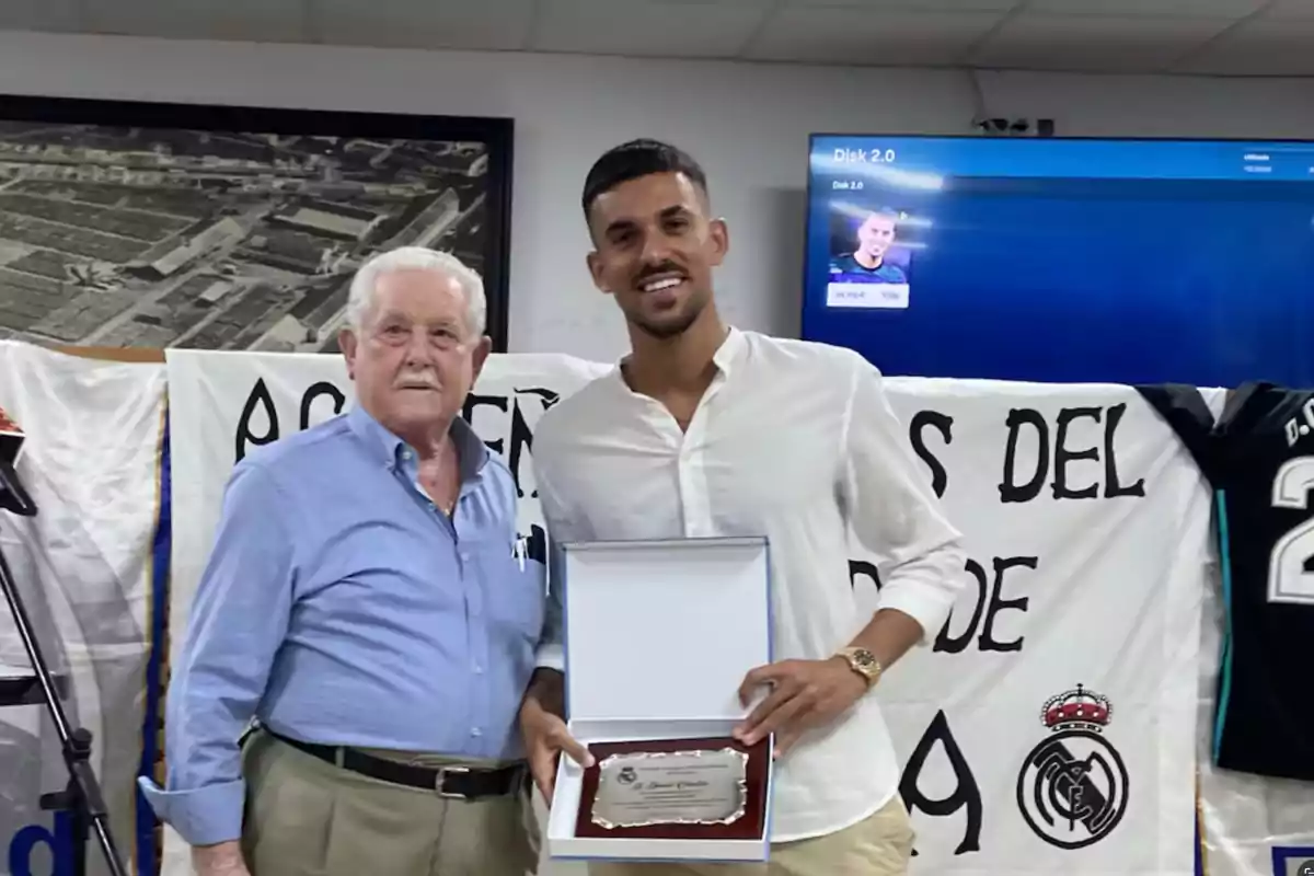 Dos hombres posan para una foto, uno de ellos sostiene una placa de reconocimiento, detrás de ellos hay una pancarta y una camiseta de fútbol.