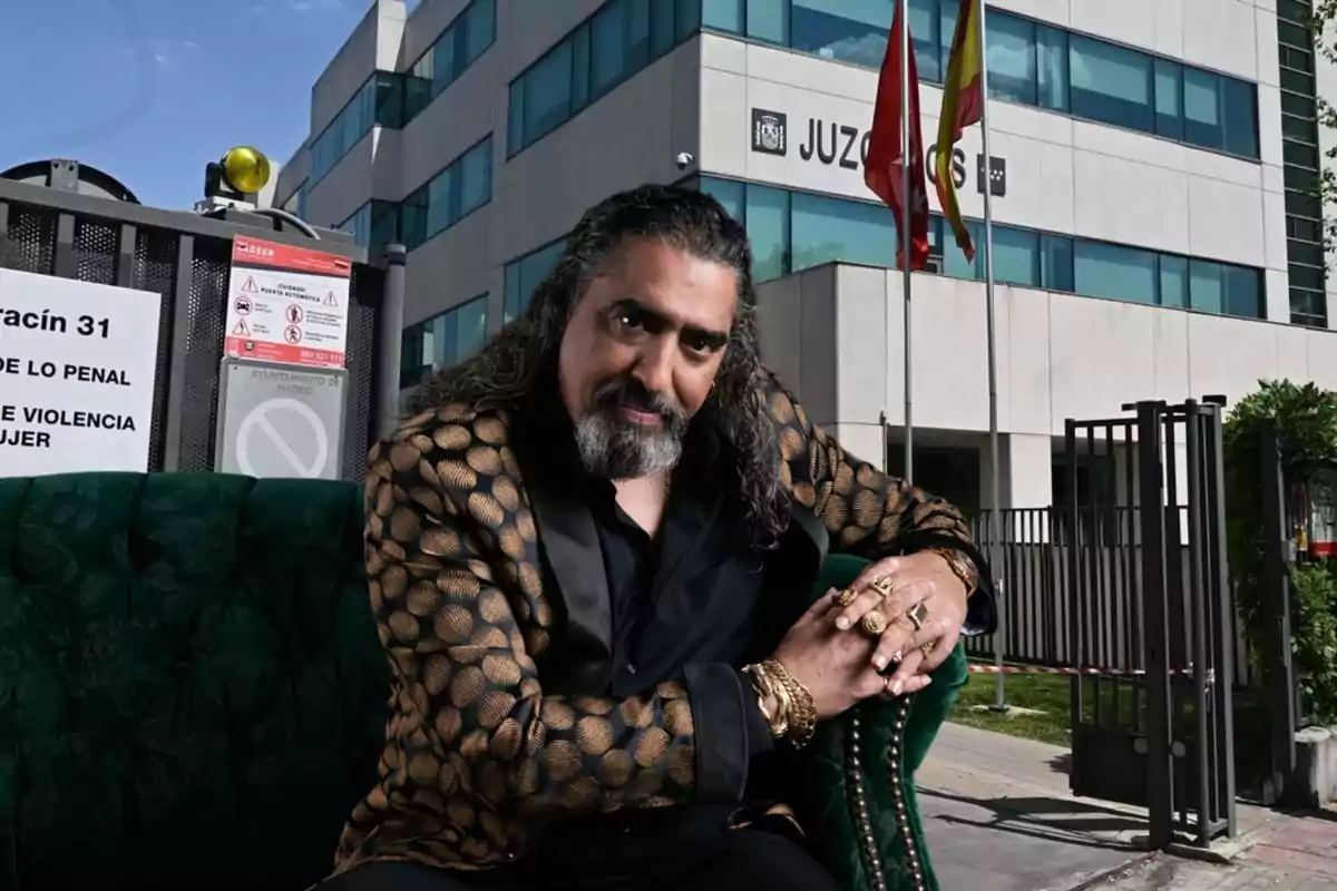 Un hombre con cabello largo y barba, vestido con una chaqueta de patrón dorado, está sentado frente a un edificio de juzgados con banderas y un cartel que indica "Juzgado de lo Penal" y "Violencia sobre la Mujer".