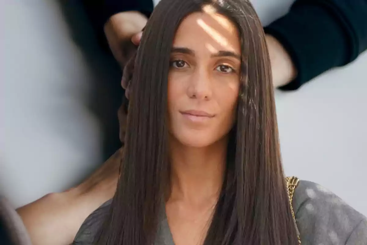 Una persona con cabello largo y liso mira directamente a la cámara mientras una mano se apoya en su hombro.