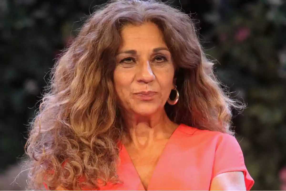 Una persona con cabello rizado y largo lleva una blusa de color coral y está al aire libre. Una persona con cabello rizado y largo lleva una blusa de color coral y está al aire libre.
