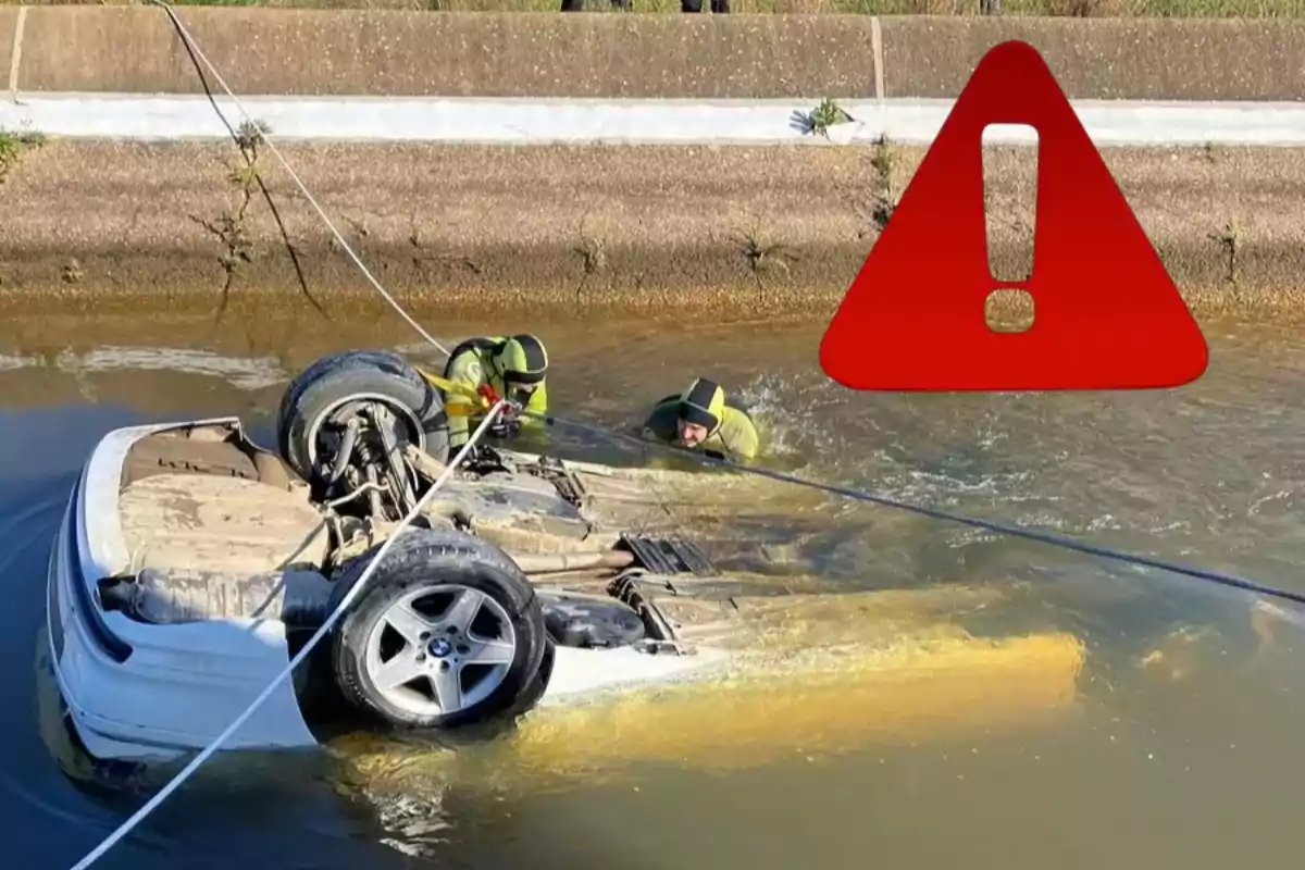 Un coche volcado en un canal de agua con dos personas en trajes de buceo trabajando para asegurar el vehículo mientras un símbolo de advertencia rojo aparece en la imagen.