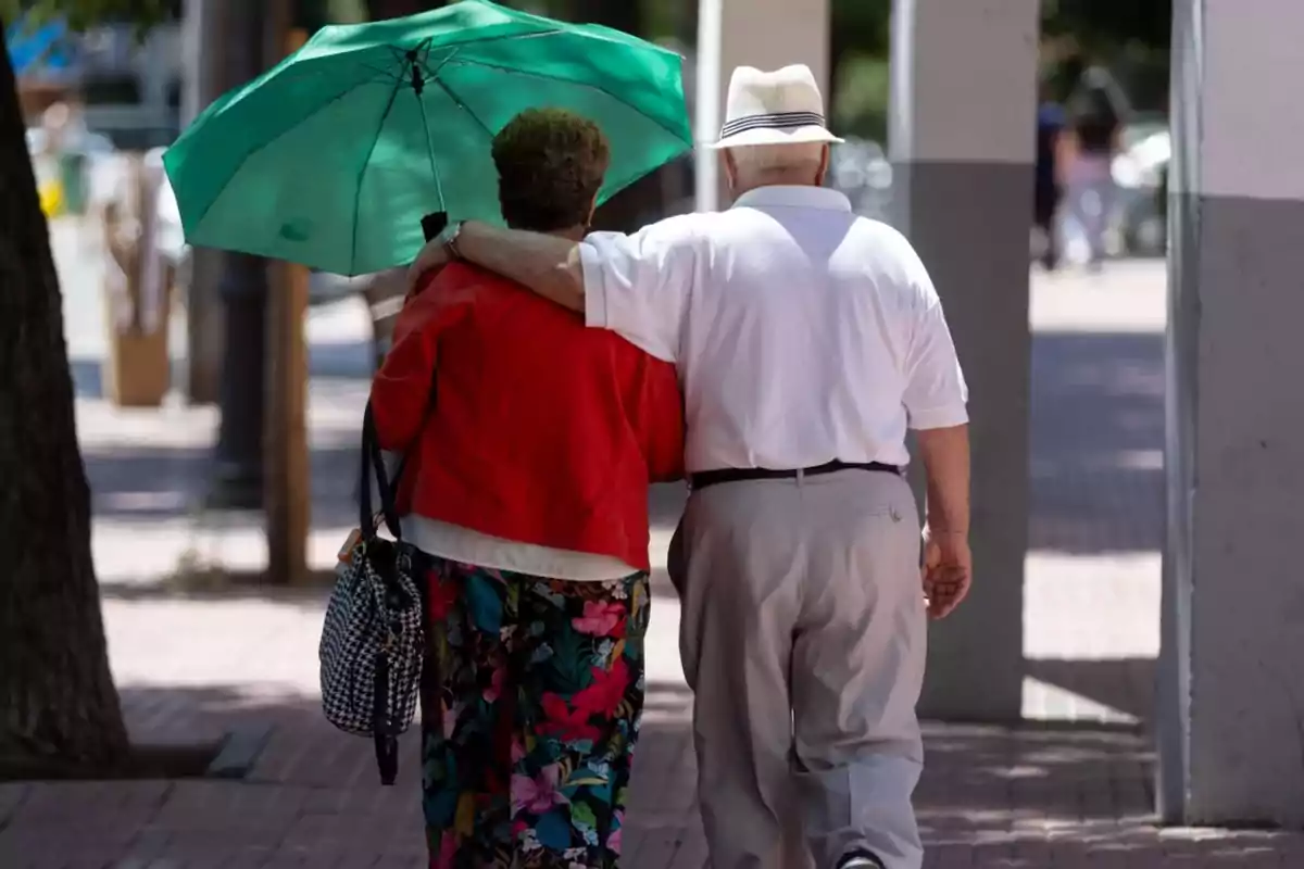 Una pareja de personas mayores caminando por la calle, el hombre lleva un sombrero y la mujer sostiene un paraguas verde.