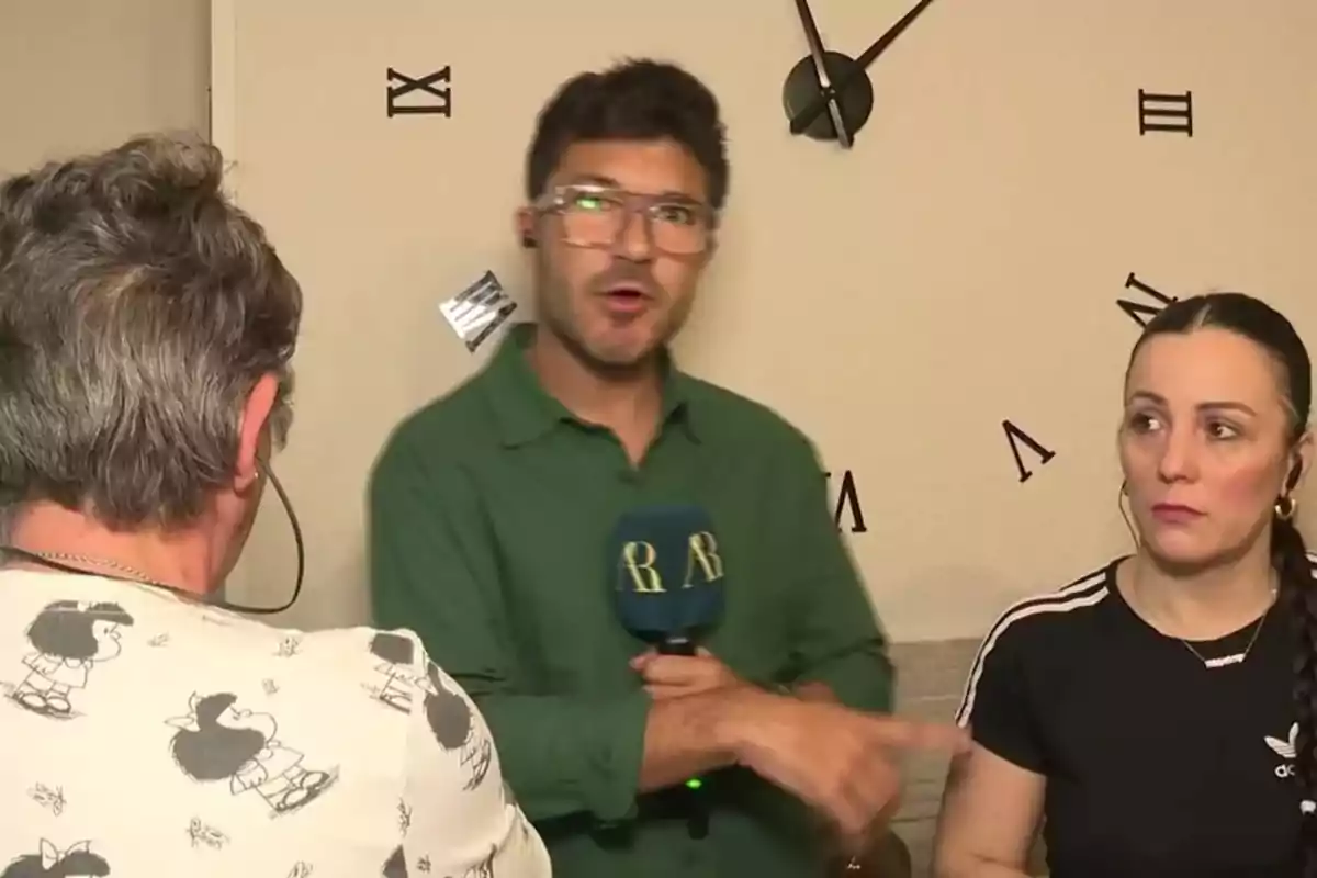 Un reportero con micrófono entrevista a dos personas en una habitación con un reloj de pared. Un reportero con micrófono entrevista a dos personas en una habitación con un reloj de pared.