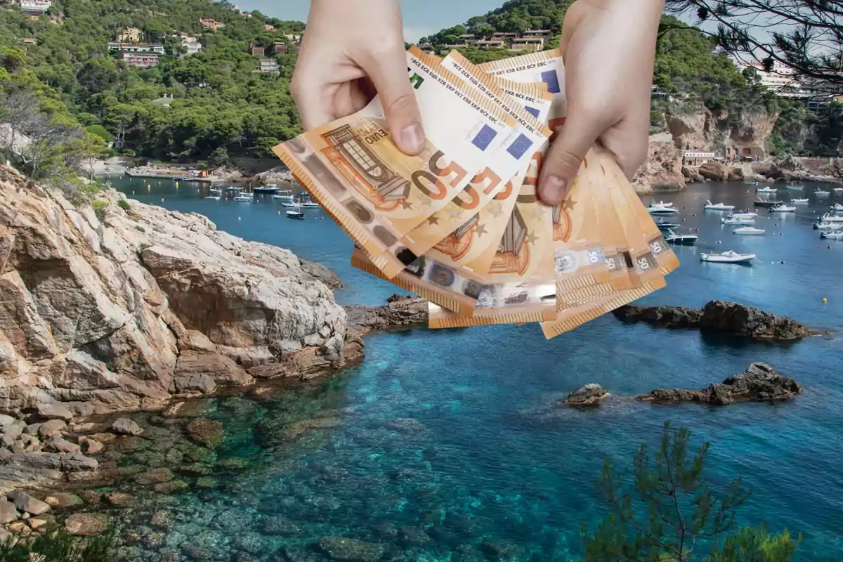 Una persona sostiene varios billetes de euro frente a un paisaje costero con agua cristalina, rocas y barcos.