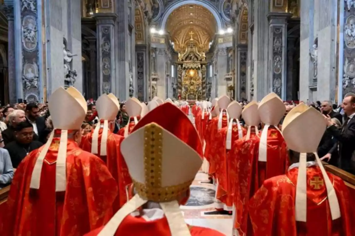 Un grupo de clérigos con vestimentas rojas y mitras blancas camina por el pasillo central de una catedral ornamentada, rodeados de fieles y bajo un techo decorado. Un grupo de clérigos con vestimentas rojas y mitras blancas camina por el pasillo central de una catedral ornamentada, rodeados de fieles y bajo un techo decorado.