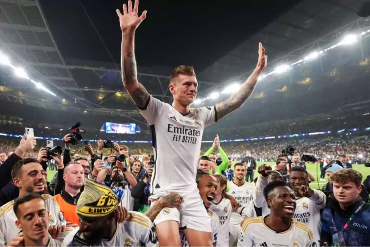 Un jugador de fútbol del Real Madrid es levantado en hombros por sus compañeros mientras celebra en un estadio lleno de aficionados y fotógrafos.