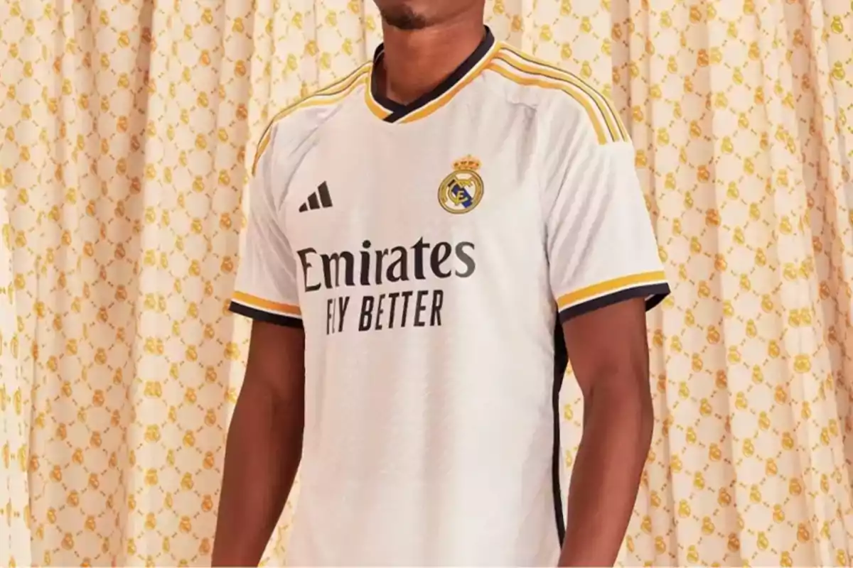 Un hombre vistiendo la camiseta blanca del Real Madrid con el logo de Adidas y el patrocinador Emirates Fly Better, con un fondo de cortinas decoradas.