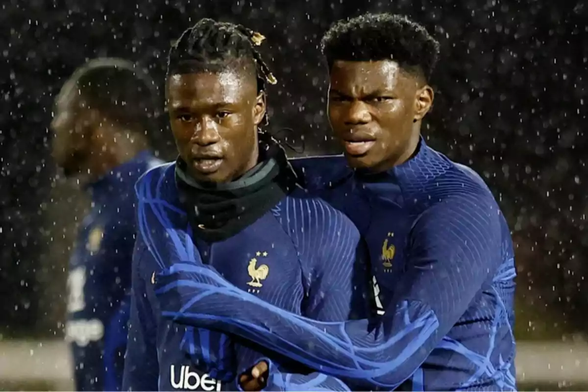 Dos jugadores de fútbol con uniformes de la selección francesa se abrazan bajo la lluvia.