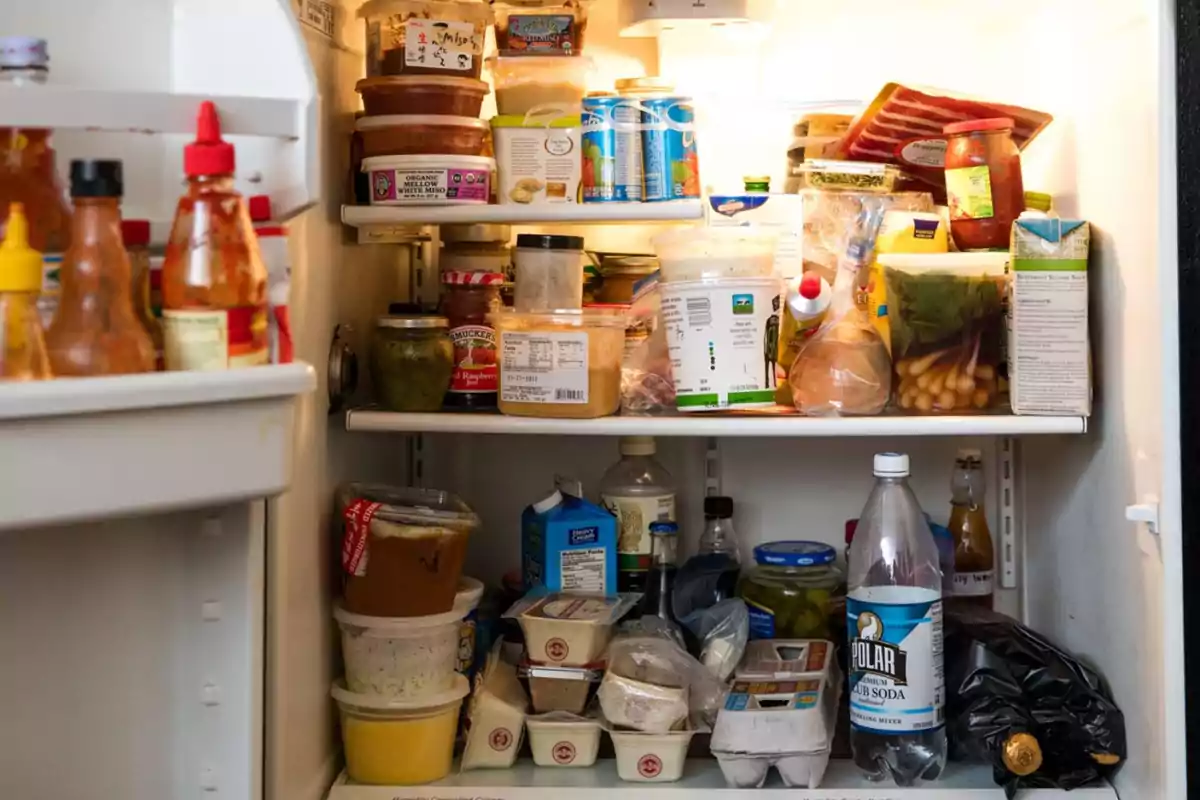 Un refrigerador lleno de diversos alimentos y bebidas, incluyendo salsas, envases de plástico, latas, botellas y productos envasados. Un refrigerador lleno de diversos alimentos y bebidas, incluyendo salsas, envases de plástico, latas, botellas y productos envasados.