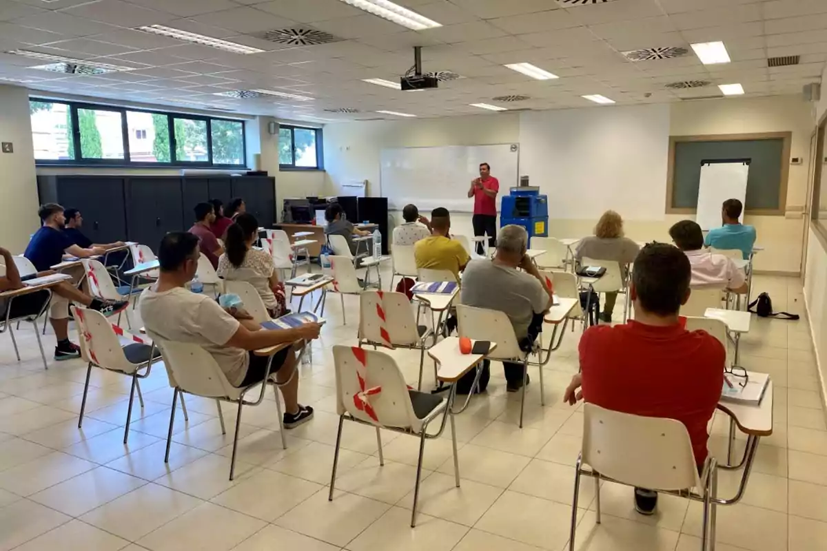 Un grupo de personas está sentado en un aula, prestando atención a un instructor que se encuentra al frente de la sala, con una pizarra blanca detrás de él.