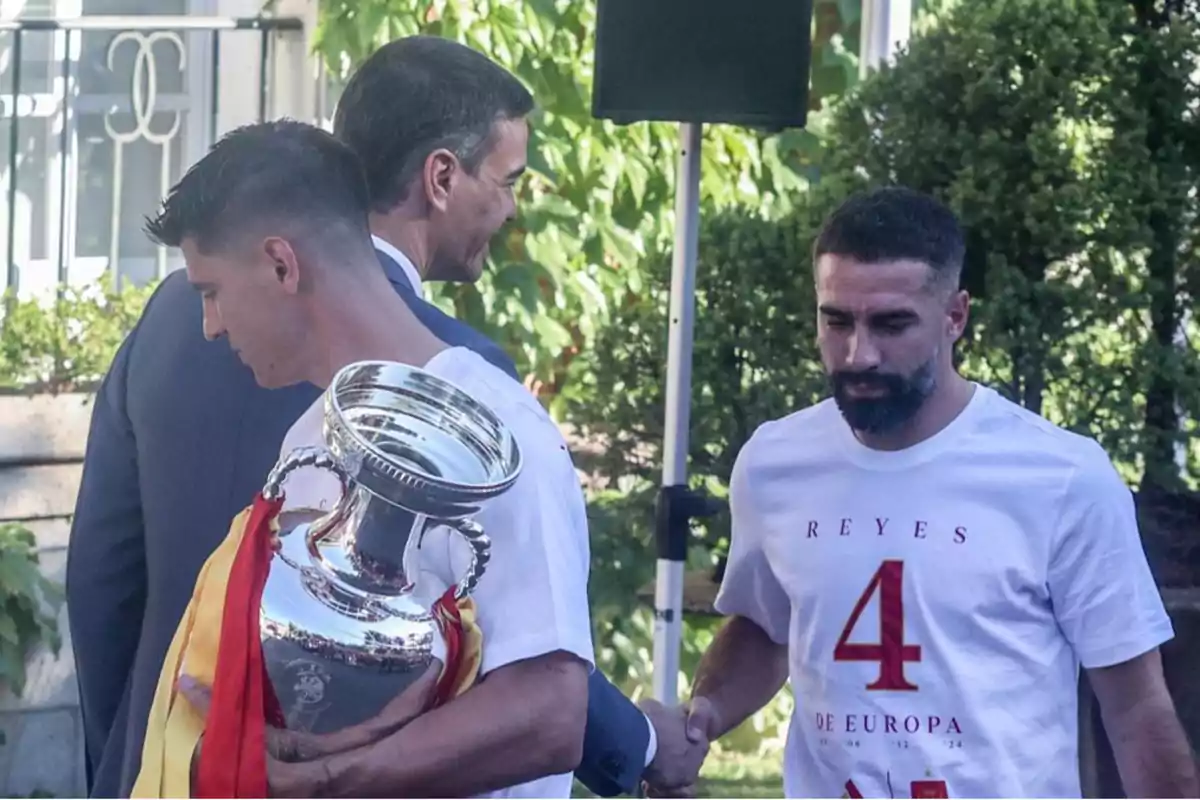 Dos hombres con camisetas blancas sostienen un trofeo plateado grande, mientras un tercer hombre con traje oscuro les da la mano en un entorno al aire libre con vegetación. Dos hombres con camisetas blancas sostienen un trofeo plateado grande, mientras un tercer hombre con traje oscuro les da la mano en un entorno al aire libre con vegetación.