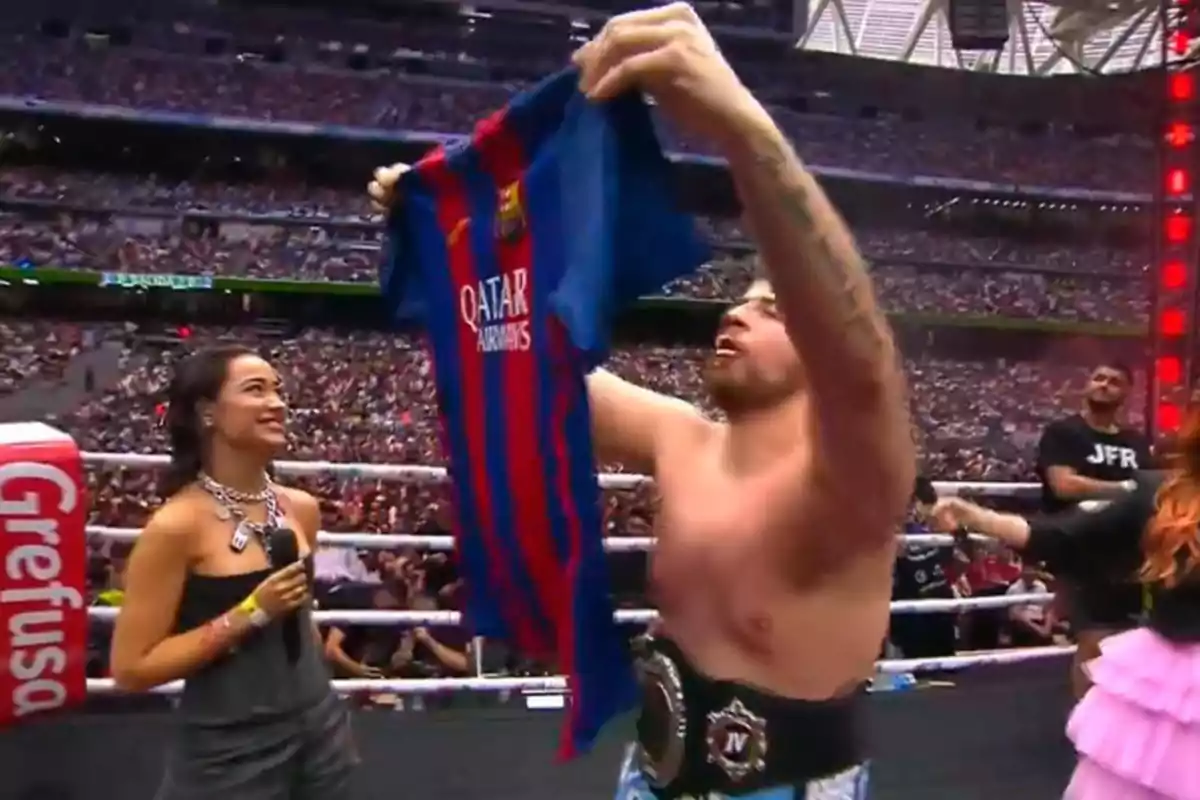 Un luchador sostiene una camiseta del FC Barcelona en un ring de lucha libre mientras una mujer con un micrófono lo observa y una multitud llena el estadio.