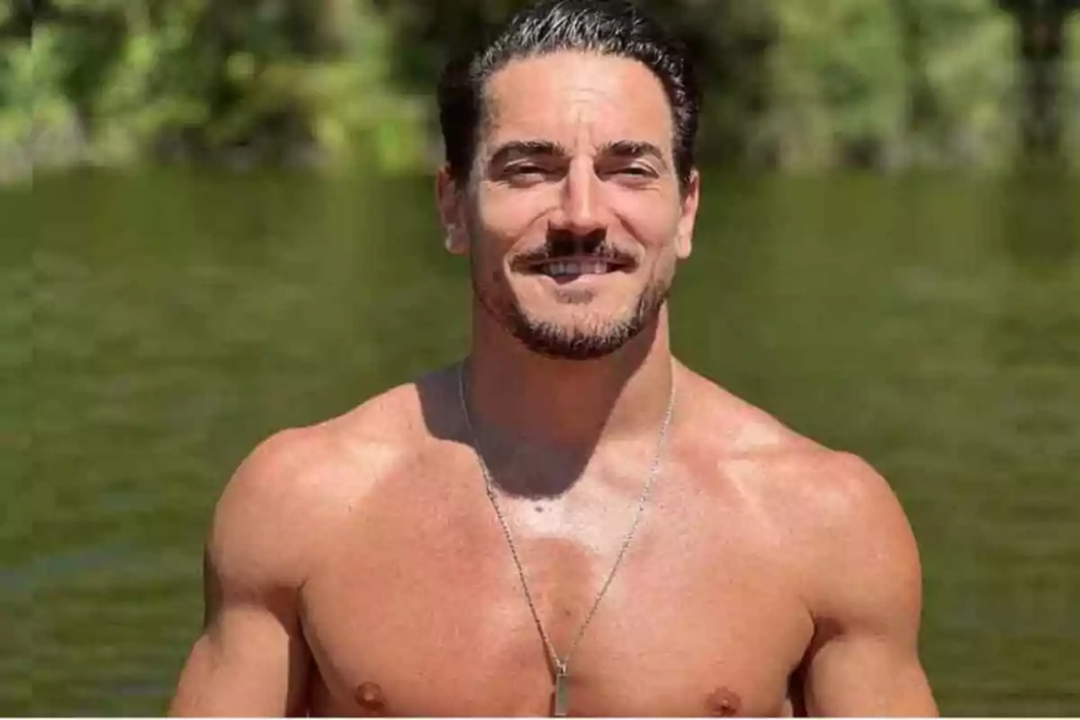 Un hombre sonriente sin camisa con un collar de cadena de pie frente a un fondo de agua y vegetación. Un hombre sonriente sin camisa con un collar de cadena de pie frente a un fondo de agua y vegetación.