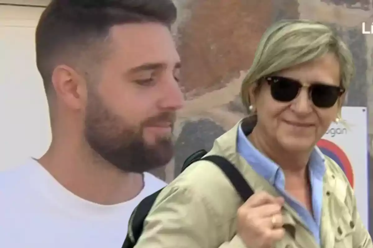 Dos personas, una con barba y camiseta blanca y otra con gafas de sol y chaqueta beige, están al aire libre frente a un fondo de piedra.