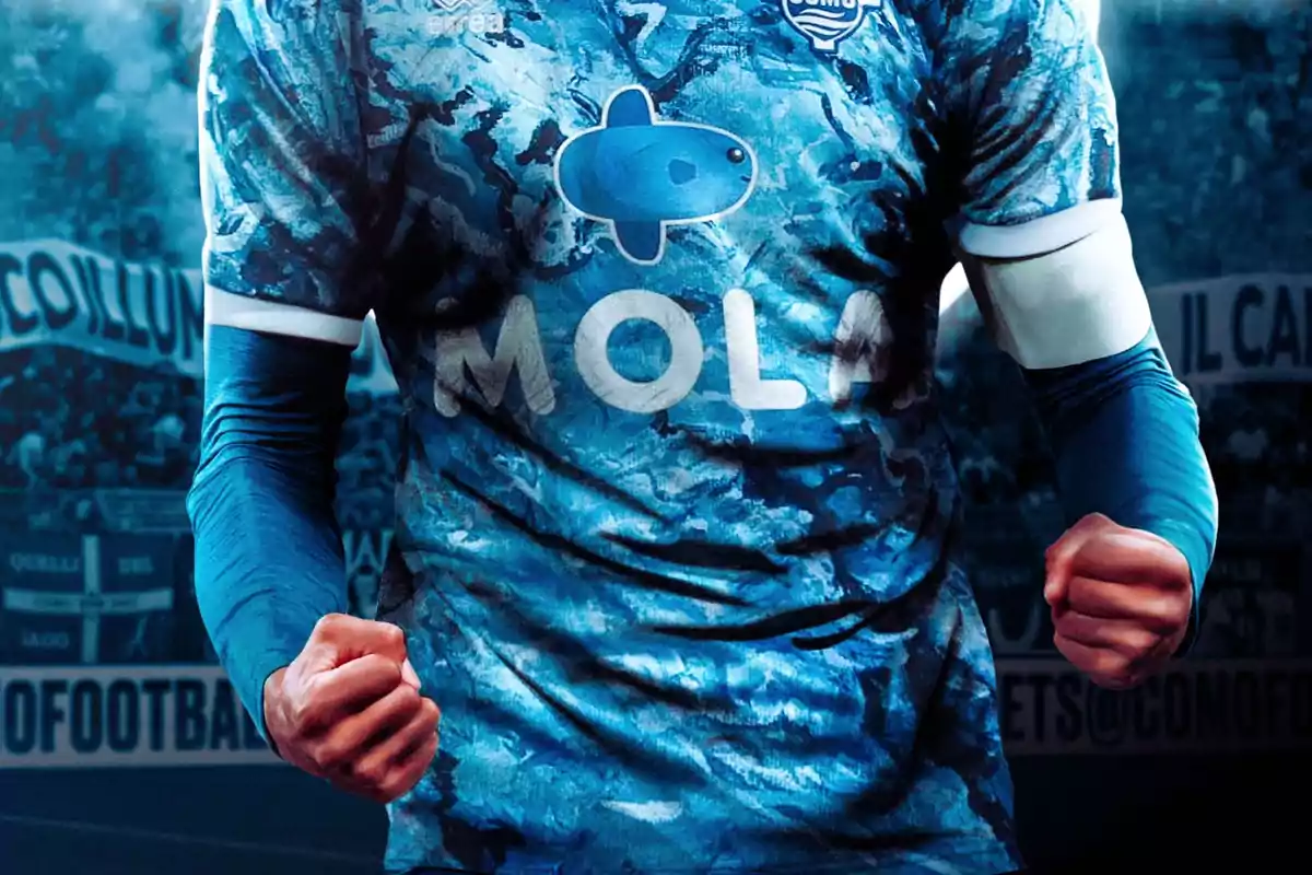 Un jugador de fútbol con una camiseta azul con un diseño de camuflaje y el logo de un pez y la palabra "MOLA" en el pecho, con los puños cerrados en una pose de celebración.