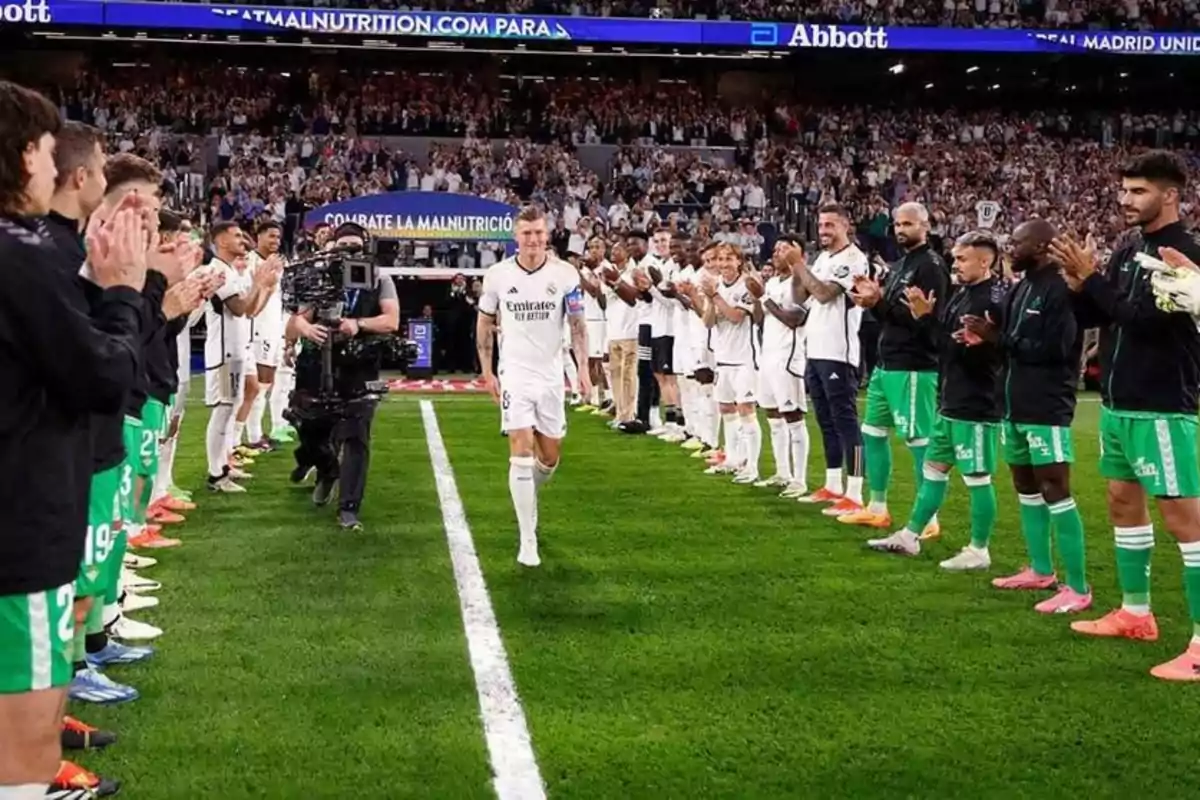 Un jugador de fútbol camina por un pasillo formado por dos filas de jugadores que lo aplauden, mientras una cámara lo graba en un estadio lleno de espectadores.