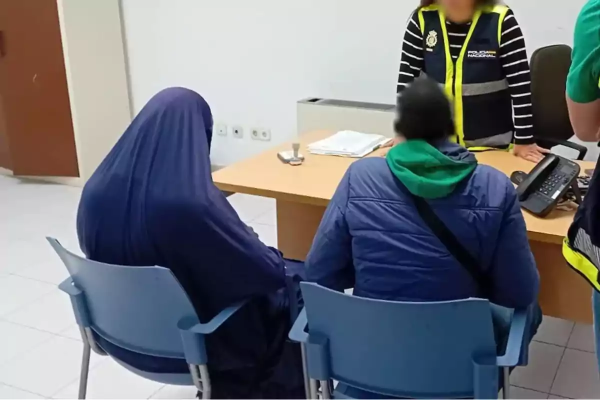 Una persona con un velo azul y otra con chaqueta azul están sentadas frente a un escritorio donde hay una persona de pie con chaleco de la policía.