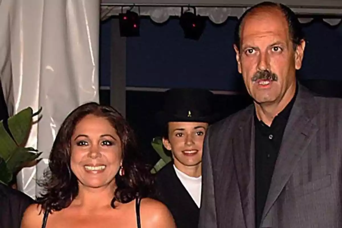 Tres personas posan juntas en un evento formal, una mujer sonriente a la izquierda, un hombre con bigote a la derecha y una persona con sombrero de fondo.