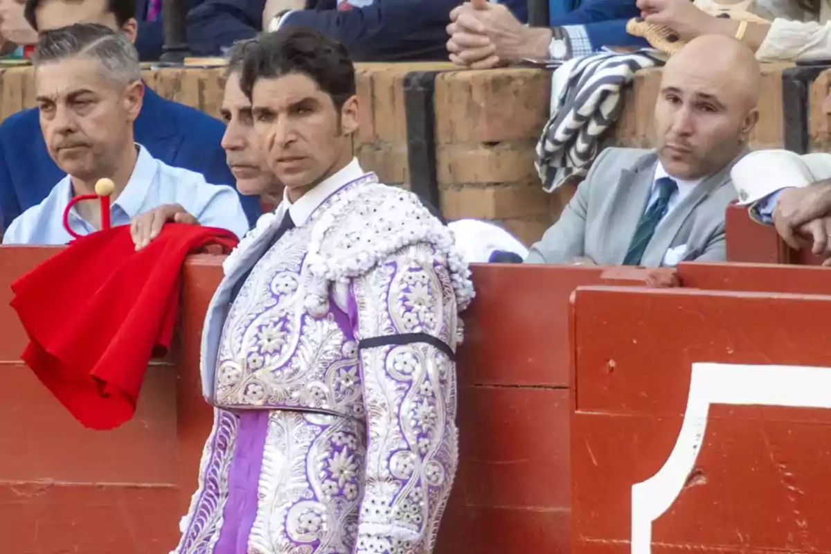 Un torero vestido con un traje de luces morado y plateado está apoyado en una barrera roja mientras otras personas observan desde el fondo. Un torero vestido con un traje de luces morado y plateado está apoyado en una barrera roja mientras otras personas observan desde el fondo.