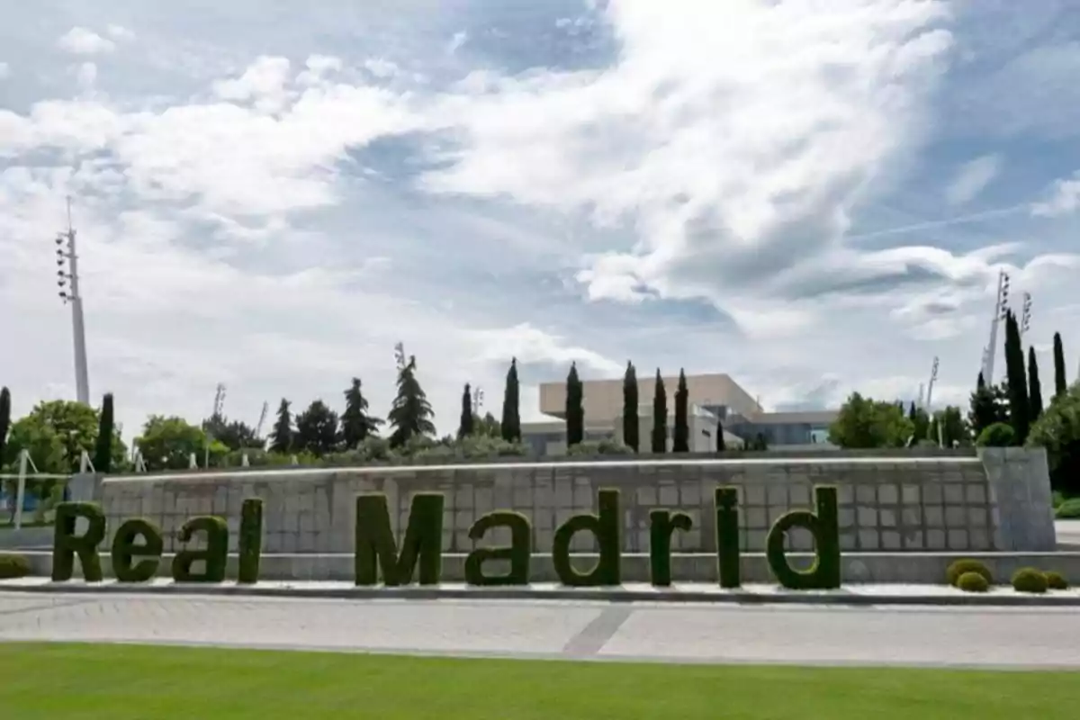 La imagen muestra un letrero de "Real Madrid" hecho de arbustos verdes frente a una estructura de concreto, con un cielo parcialmente nublado y árboles en el fondo.