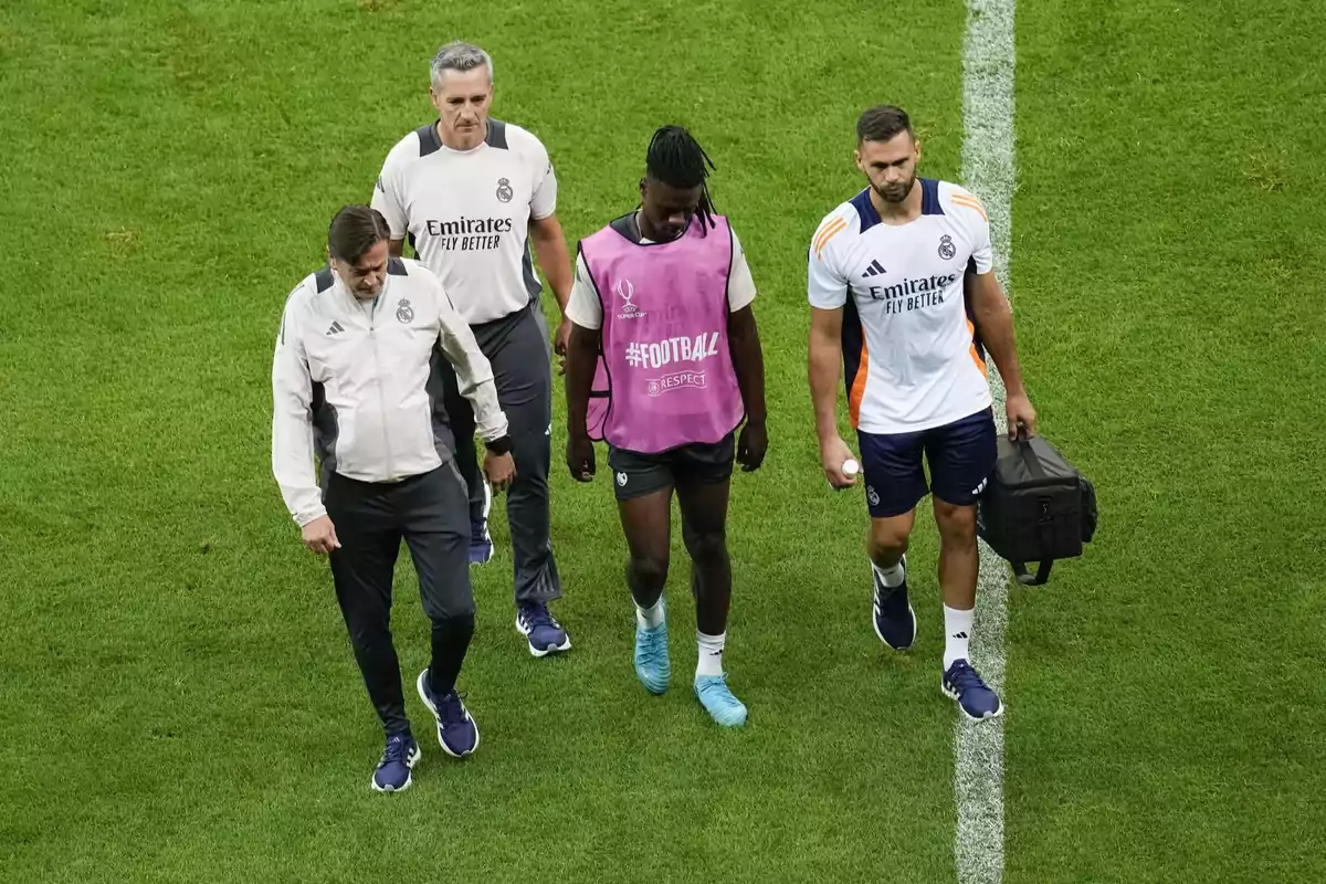 Un jugador de fútbol camina acompañado por tres miembros del personal del equipo en un campo de fútbol.