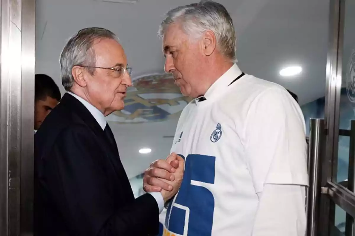 Dos hombres se saludan estrechando las manos, uno de ellos lleva un traje oscuro y el otro una camiseta blanca con el logo de un equipo de fútbol.