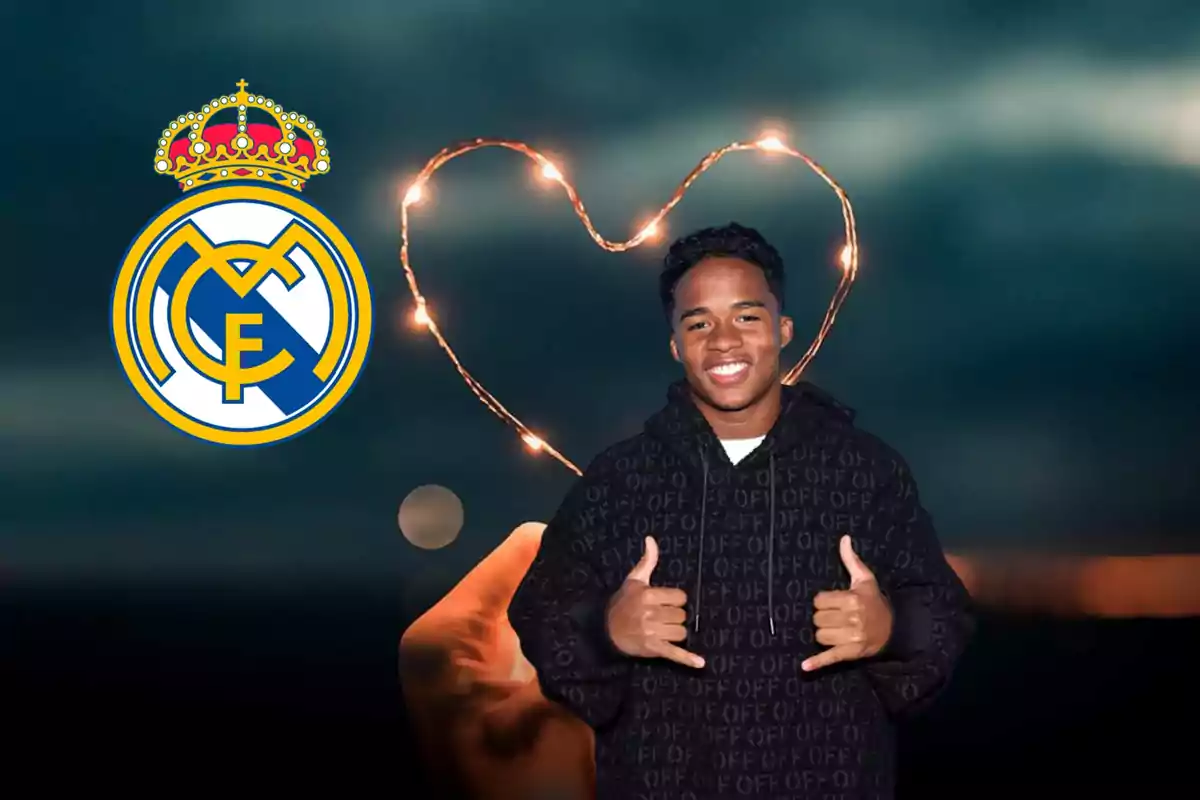 Un joven sonriente con una sudadera negra hace un gesto de aprobación con ambas manos, mientras que a su izquierda se encuentra el escudo del Real Madrid y al fondo se ve una mano sosteniendo luces en forma de corazón.