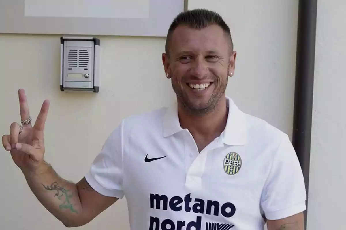 Un hombre sonriente con una camiseta blanca del Hellas Verona hace el signo de la paz con la mano.