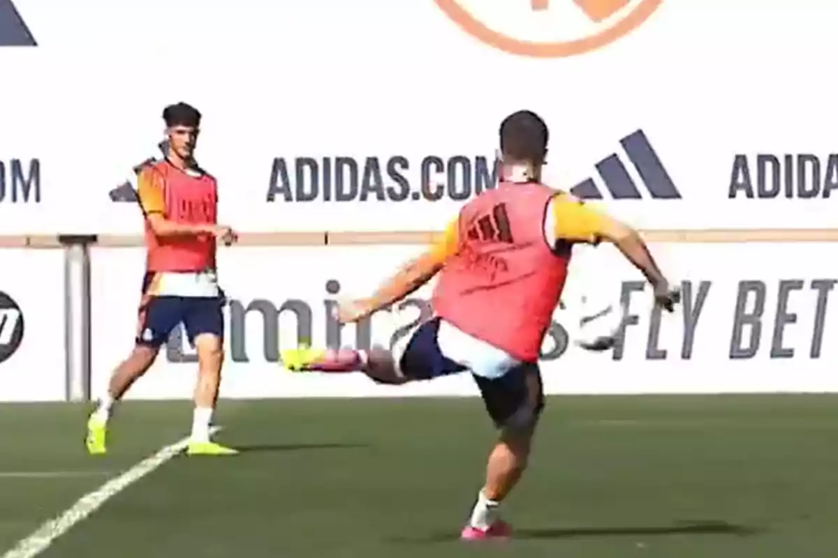 Dos jugadores de fútbol entrenan en un campo con petos rojos y camisetas amarillas, uno de ellos está pateando el balón mientras el otro observa.