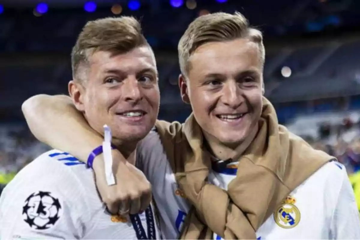 Dos hombres sonrientes con camisetas del Real Madrid, uno de ellos con una medalla y el otro con un suéter beige sobre los hombros.