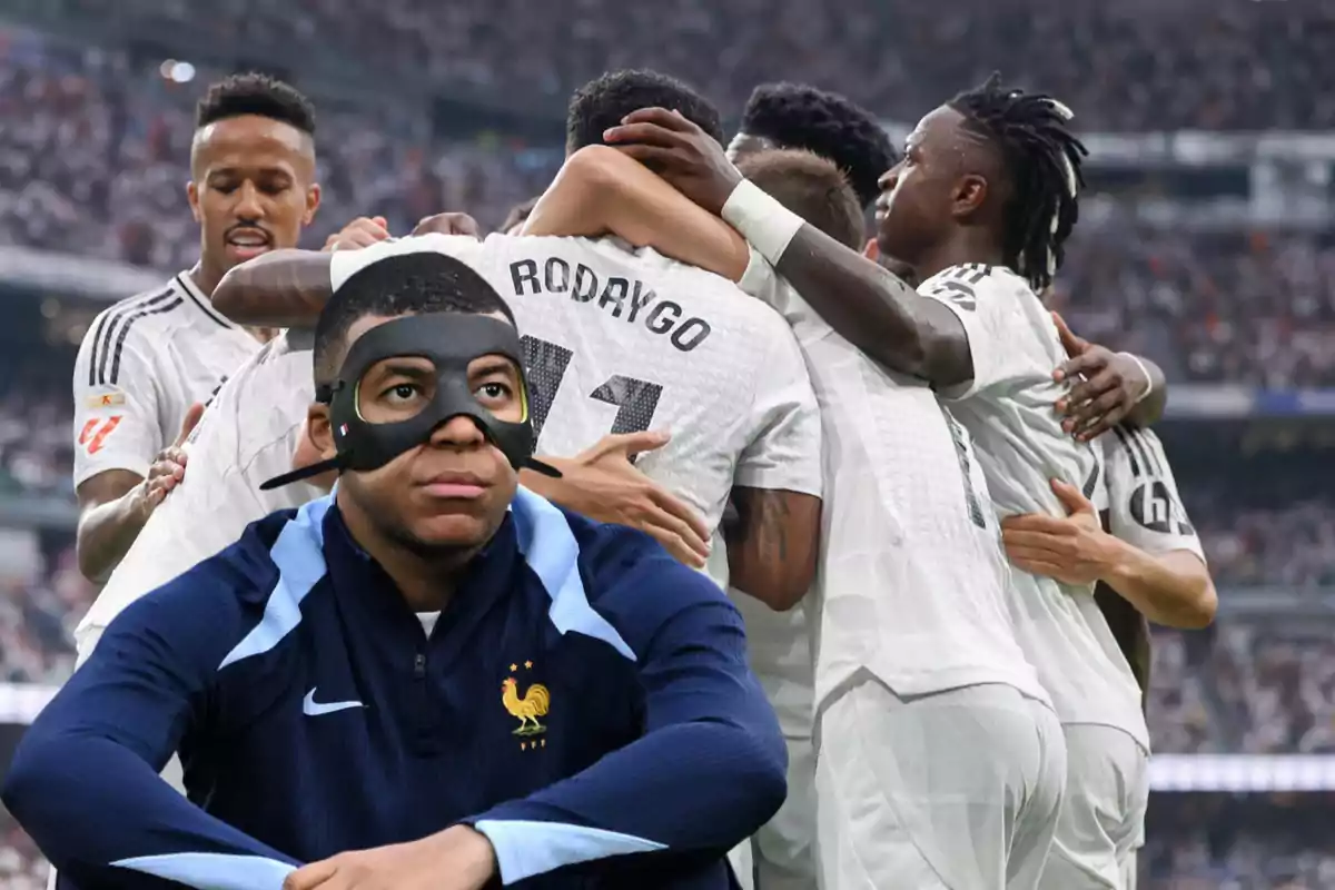 Un jugador con máscara negra y uniforme azul oscuro está sentado en primer plano mientras un grupo de jugadores de fútbol con uniformes blancos se abrazan celebrando en el fondo.