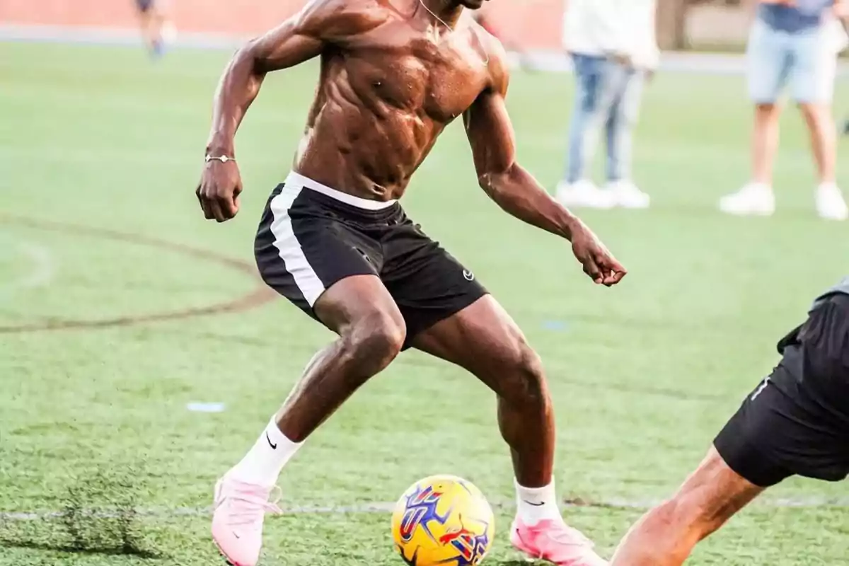 Un hombre musculoso jugando al fútbol en un campo de césped, vistiendo pantalones cortos negros y zapatillas rosas.